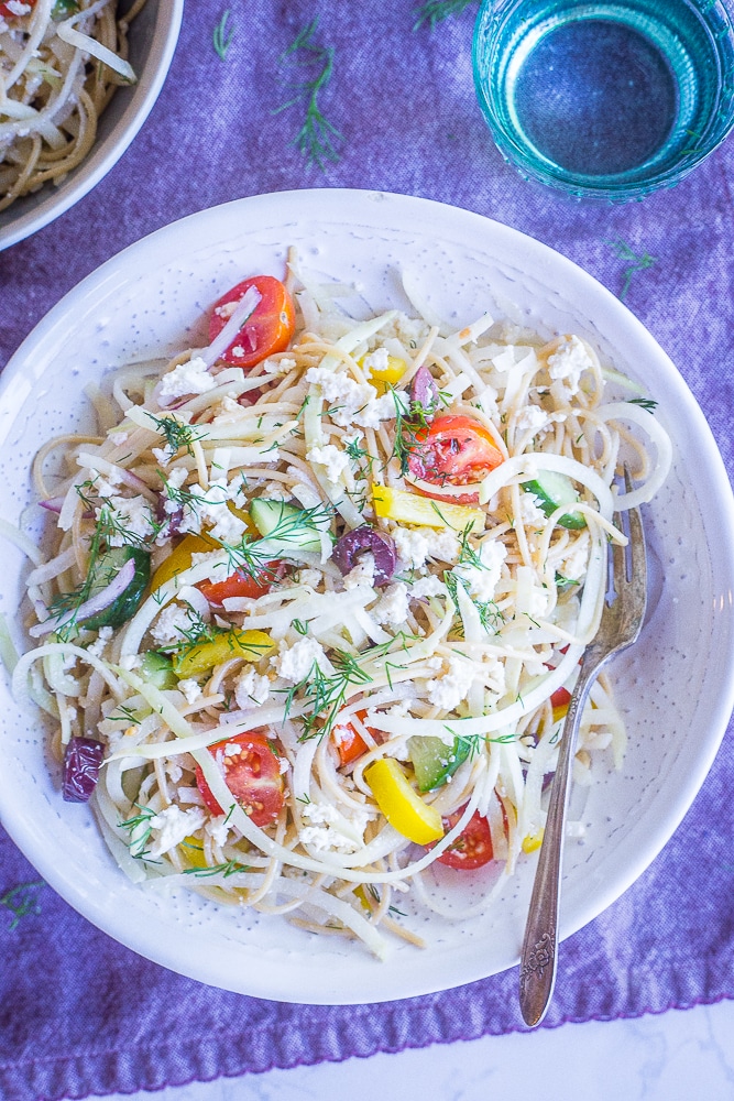 A bowl of Healthy Greek Pasta Salad with Kohlrabi Noodles in a white bowl on a purple napkin