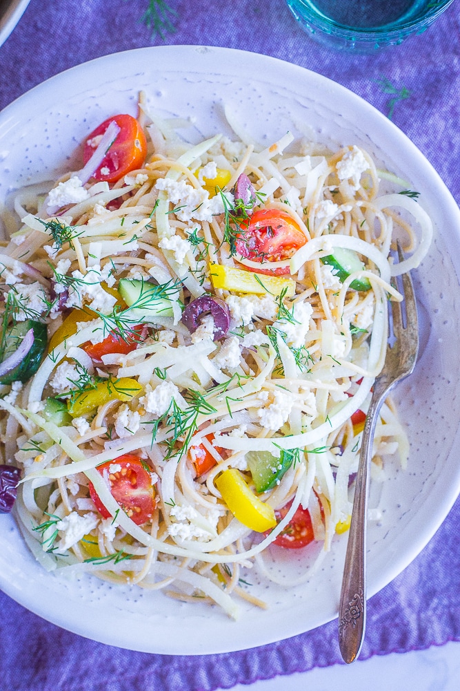 A bowl of Healthy Greek Pasta Salad with Kohlrabi Noodles in a white bowl on a purple napkin