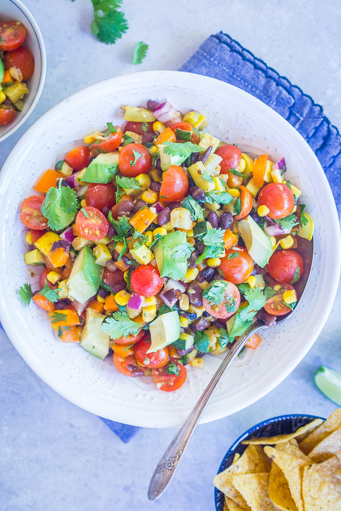 A big bowl of Loaded Summer Vegetable Salad with Black Beans on a blue napkin with a side of chips.