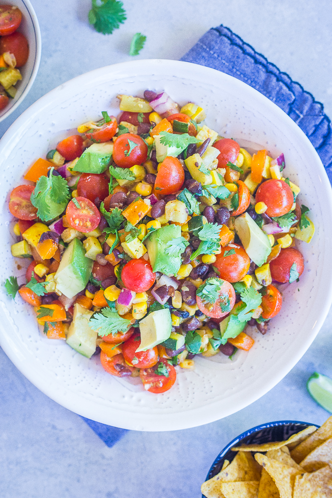 A big bowl of Loaded Summer Vegetable Salad with Black Beans on a blue napkin with a side of chips.