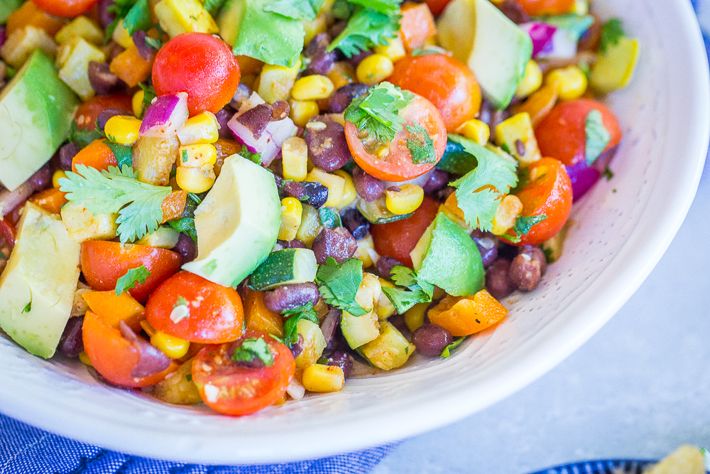 A big bowl of Loaded Summer Vegetable Salad with Black Beans on a blue napkin with a side of chips.