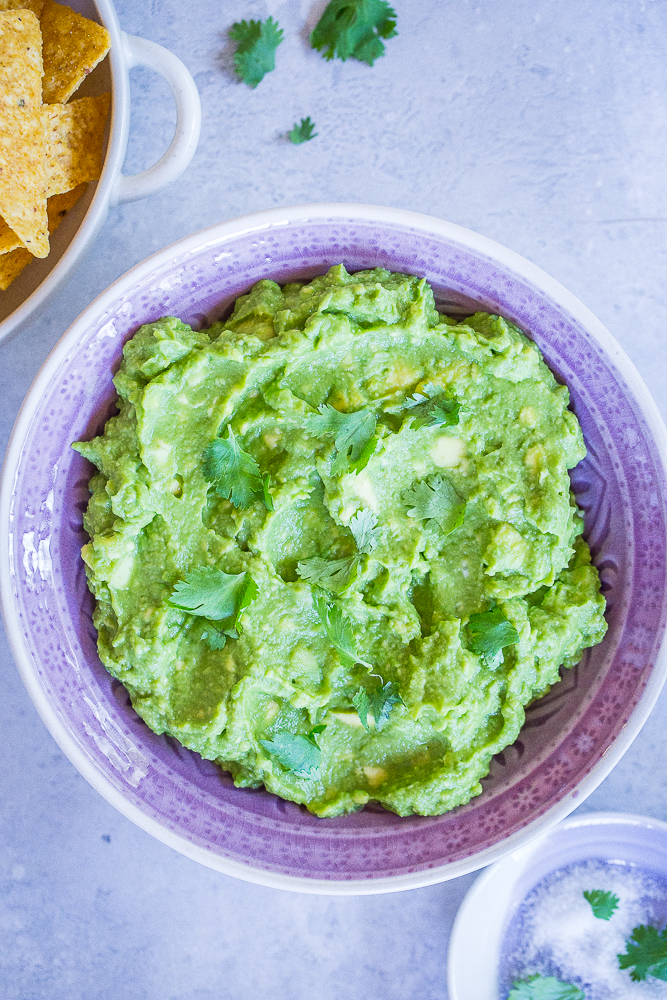 A bowl of The Best Easy Guacamole Recipe in a purple bowl with some chips and salt.