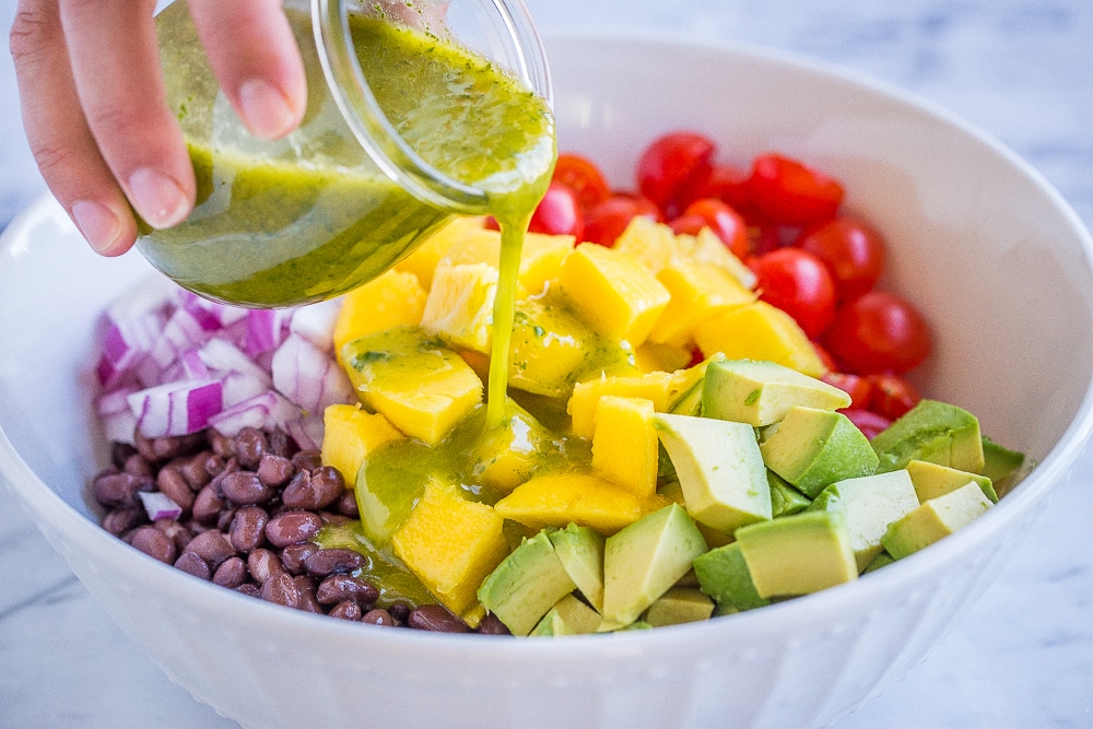 Hand pouring a glass of dressing over the black bean salad with mango and avocado