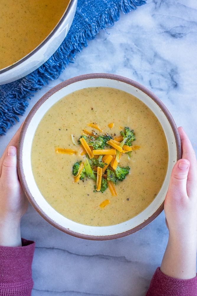 hands holding a bowl of creamy broccoli cheddar soup