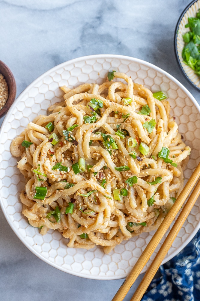 ginger tahini noodles in a bowl with a side of fresh herbs and toasted sesame seeds