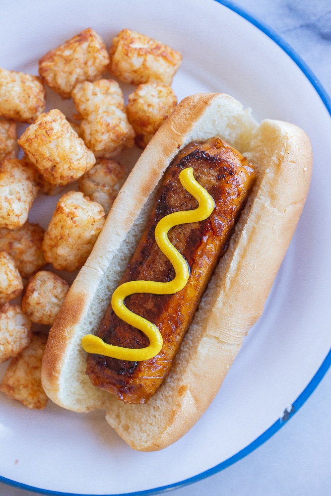vegan hot dog in a bun topped with mustard and tater tots