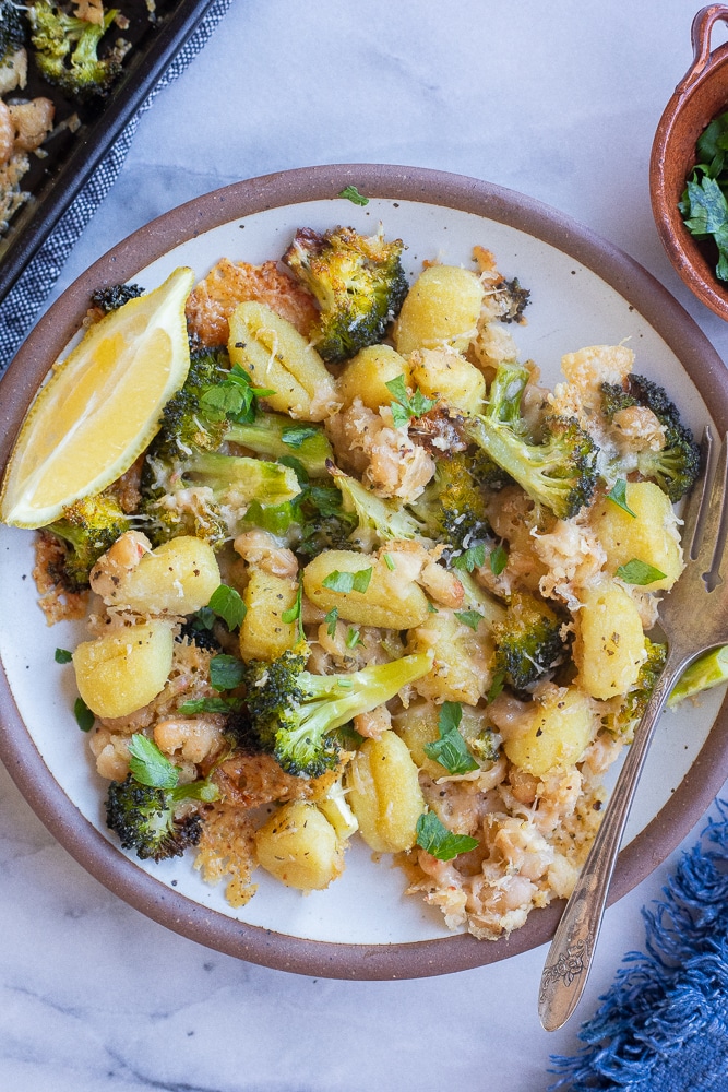 plate full of parmesan crusted gnocchi with broccoli and white beans with a lemon wedge and fork