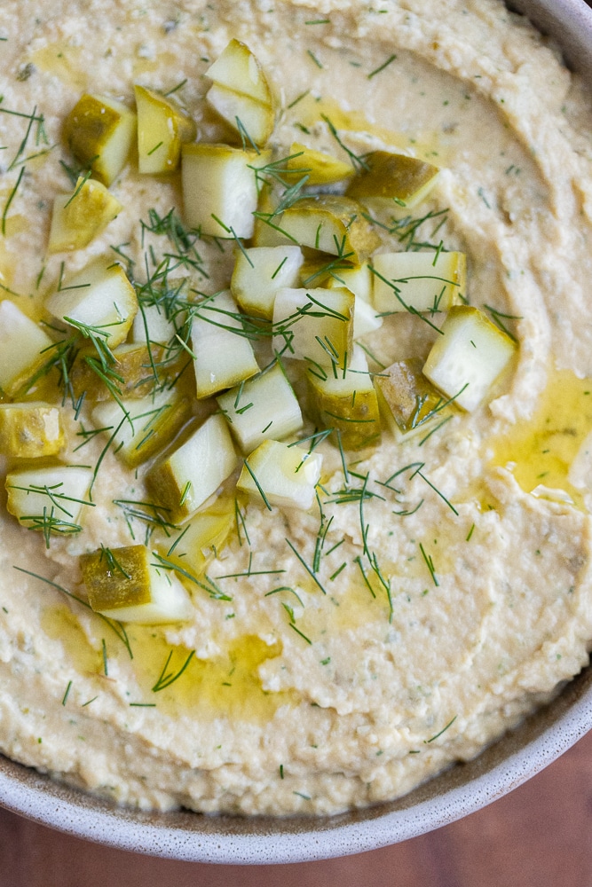 close up of a bowl of dill pickle hummus topped with pickles, fresh dill and olive oil