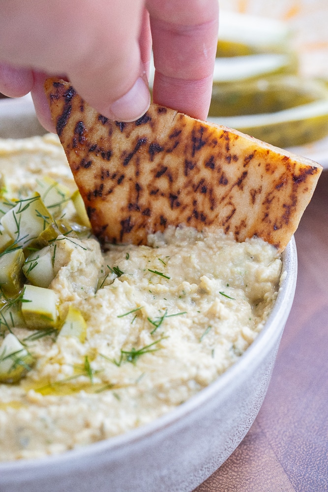 a hand dipping a piece of pita bread in a bowl of creamy dill pickle hummus